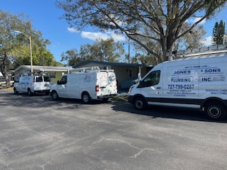 Three white plumbing service vans line a residential street in front of a single-story house surrounded by trees.